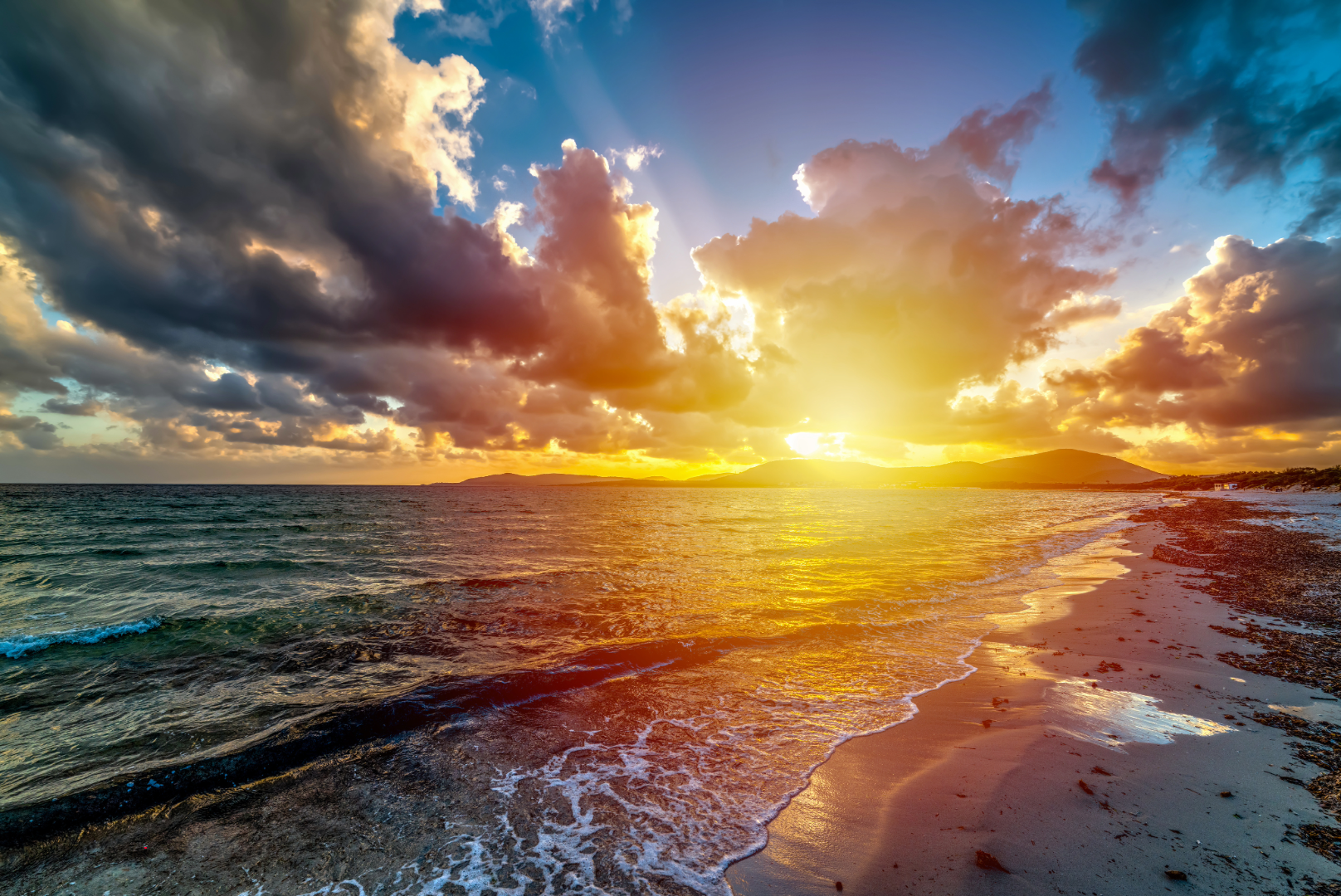 Blue and Gold sunset from a beach with waves and hills in the distance.